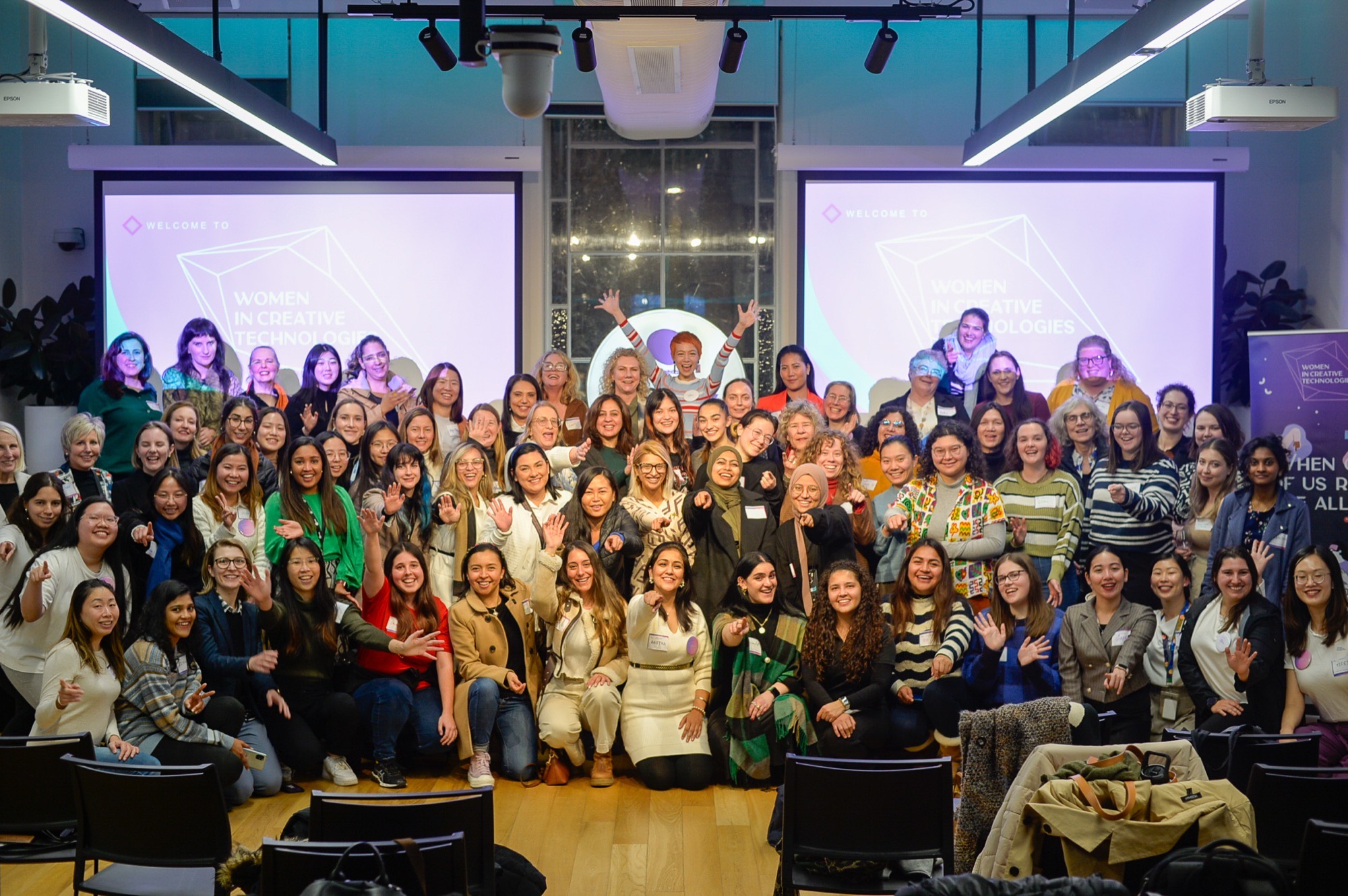 A large group of women at the WiCT launch night at Stone and Chalk, waving at the camera in front of screens reading Welcome to Women in Creative Technologies.